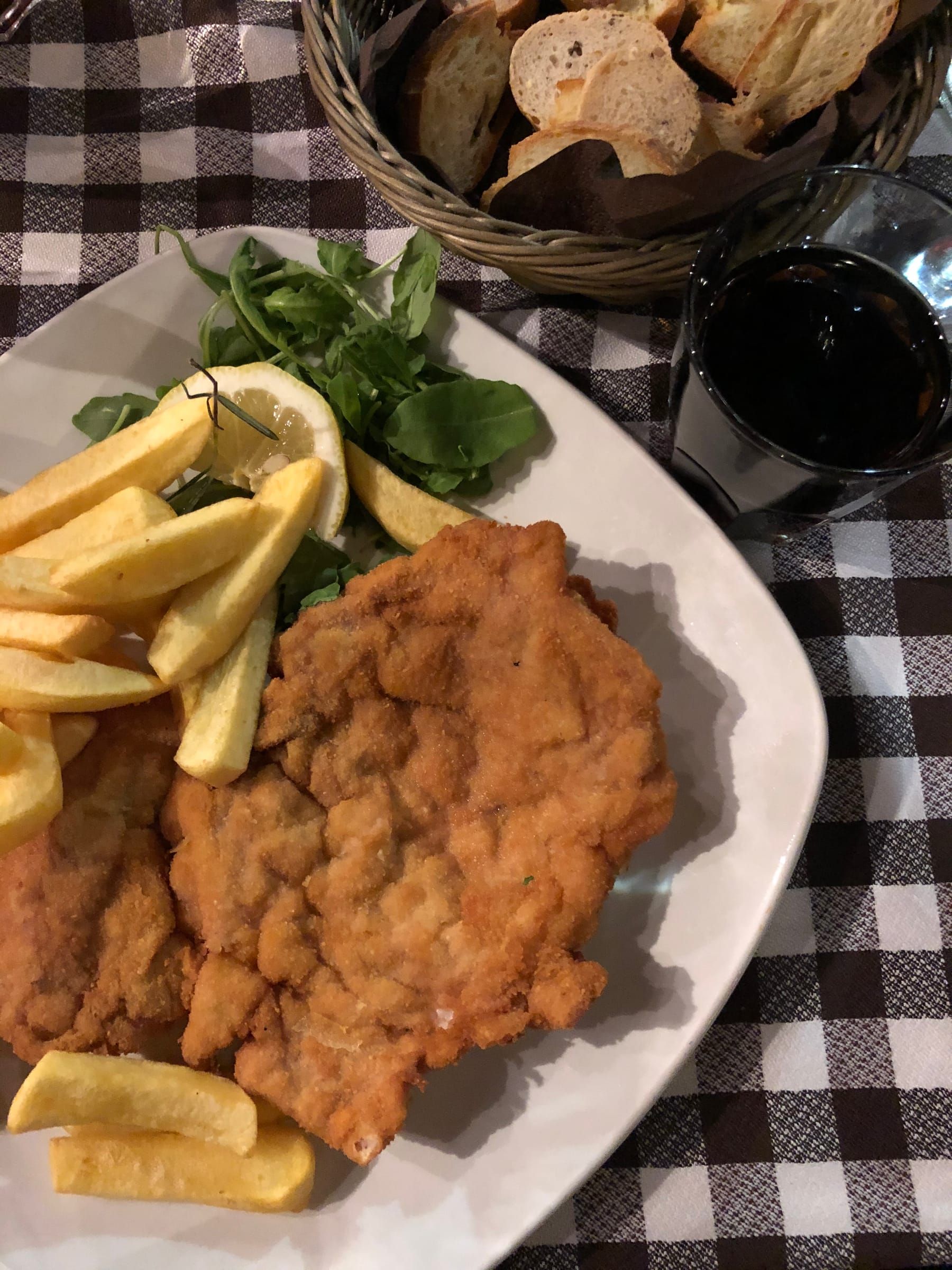 A cotoletta with fries, a basket of bread and a glass of red wine on the checked tablecloth of a trattoria