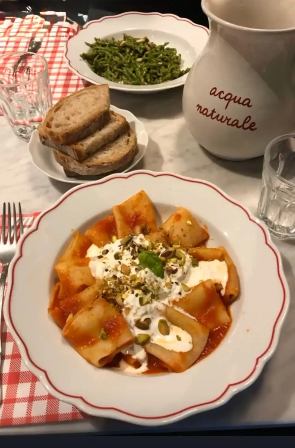 A plate of paccheri with tomato, stracciatella and pistachios, with bread and a water jug beside it