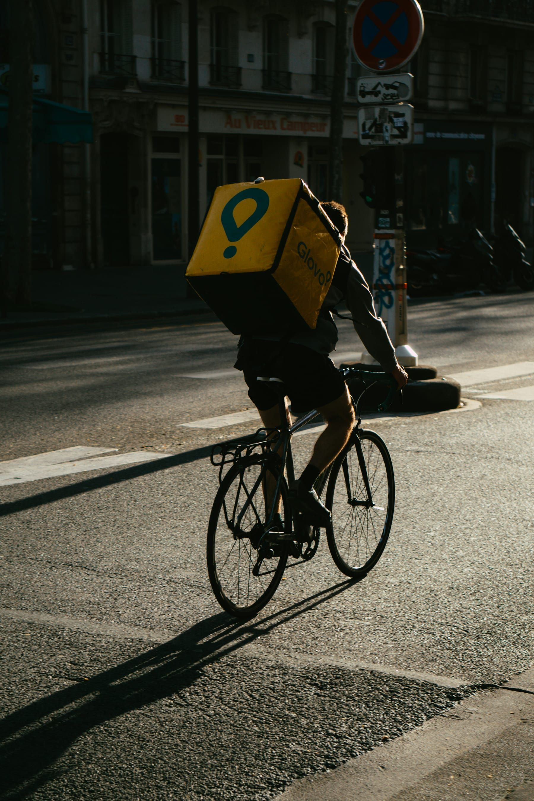 A rider on a bicycle with a yellow thermal backpack crosses a Milan street at sunset